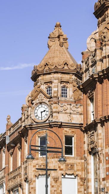 A tall, historic building with ornate architectural details made of red brick and sandstone, featuring a rounded clock tower with a large white clock face displaying black Roman numerals and hands, set against a clear blue sky. The building has small arched windows and decorative stonework, with a conical tiled roof topped by a decorative finial. In the foreground, a street lamp with two round light fixtures is visible, mounted on a black pole. This scene is typical of urban architecture often encountered during house removals or relocation services in Croydon, such as those provided by Removal Company Croydon, supporting the context of the Croydon removals guide for North End and George Street.