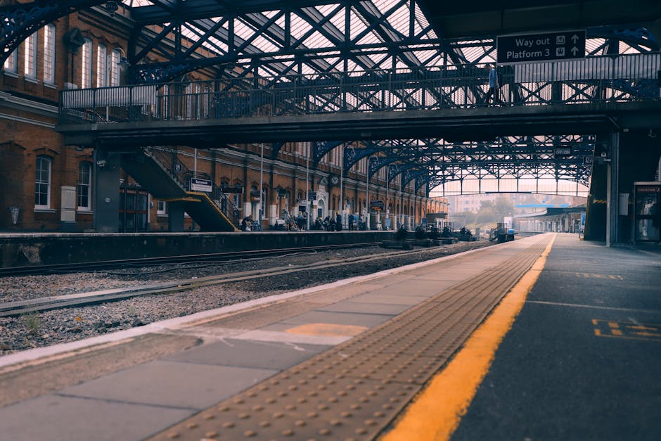 View of East Croydon railway station platform with a covered metal and glass canopy overhead, showing train tracks running parallel to the platform. On the platform, several cardboard boxes and packaged furniture covered in protective blankets are being carried or prepared for loading onto a removal company's van. A nearby trolley is visible, assisting with the transportation of items. The scene captures the loading process during a house removal, with staff from Removal Company Croydon coordinating furniture transport and packing materials in a well-lit, urban environment, highlighting the logistical aspects of home relocation and moving services.