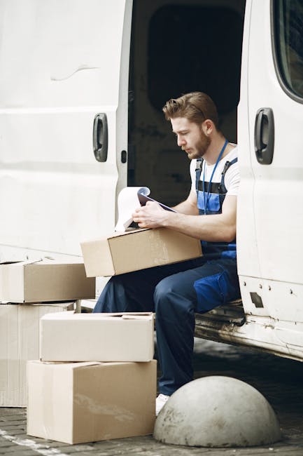 A young male mover with short, styled hair and a trimmed beard, dressed in blue work overalls, is seated at the rear entrance of a white moving van. He is holding a clipboard or checklist in one hand 
